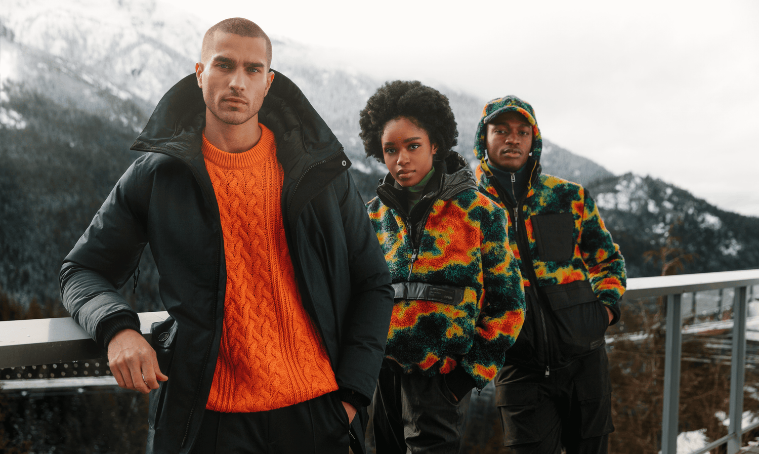 Three models lean on a railing with snowy mountains in the background. From left to right, models wear: Nobis Yves, Nobis Roche, Nobis Kepler.